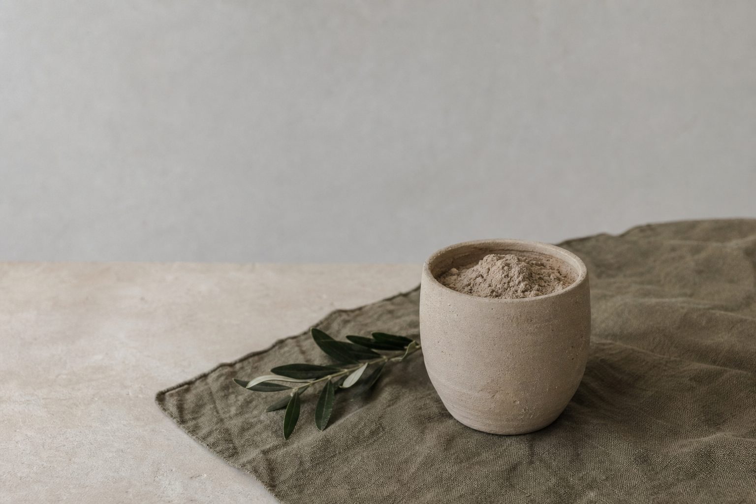 A plain ceramic vessel of pale soil and a green sprig on an ivory table in soft grey light