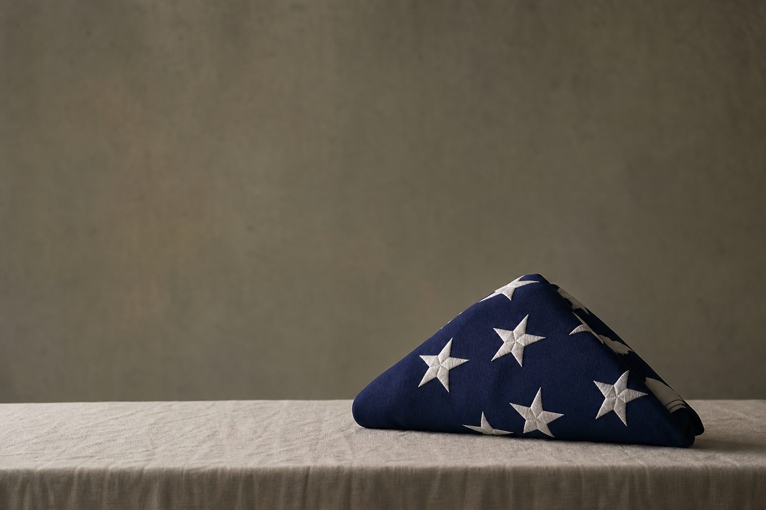 A folded United States burial flag resting on ivory linen in soft grey light against an olive wall