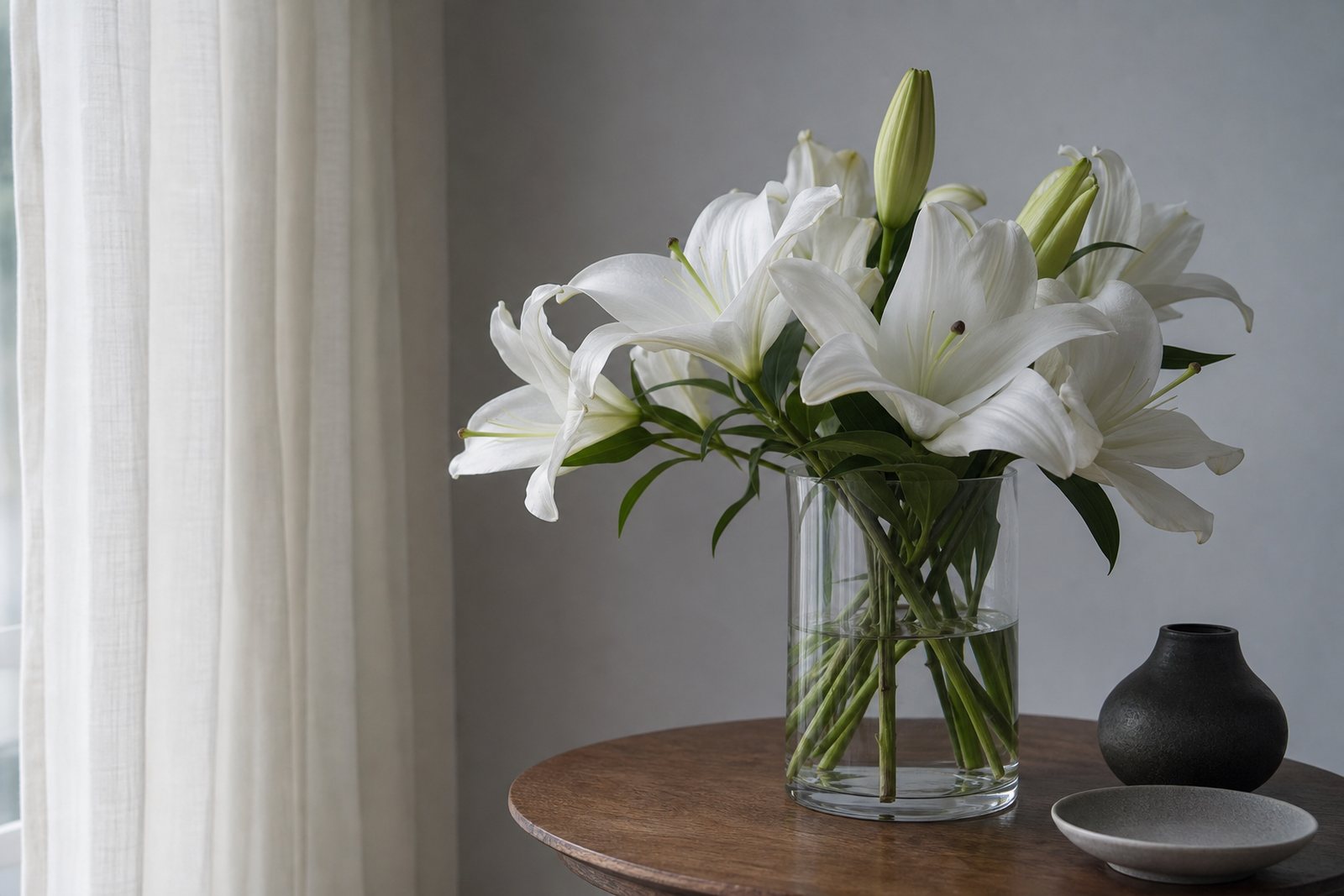 White lilies in a clear glass vase on a wooden side table in soft window light